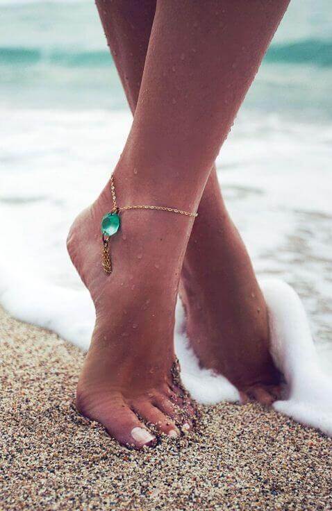 Delicate gold anklet with turquoise pendant on bare feet in shallow ocean water, highlighting boho style and beach aesthetics.