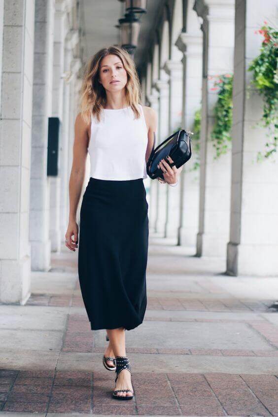 Sleeveless white top paired with a mid-length black skirt, strappy black sandals, and a structured black bag against elegant stone columns.