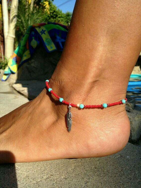 Anklet with red and turquoise beads and a silver feather charm on a tanned ankle, set against a tropical pool background.