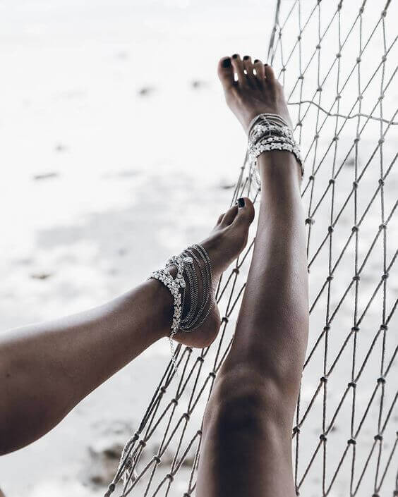 Ankle bracelets adorn two tanned legs on a sandy backdrop, featuring intricate silver chains that enhance a relaxed, beachy vibe.