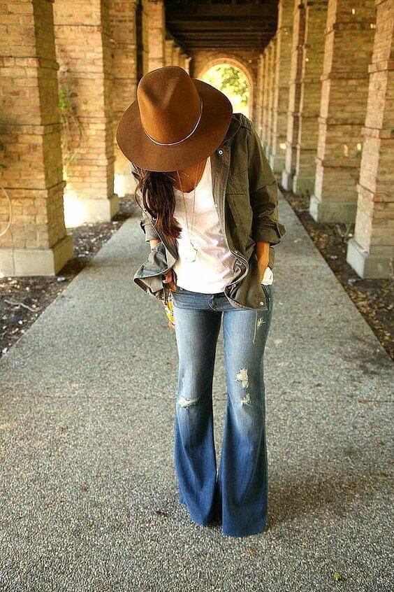 Casual outfit with distressed flared jeans, a light cotton t-shirt, brown felt hat, and olive green jacket, perfect for mild weather.