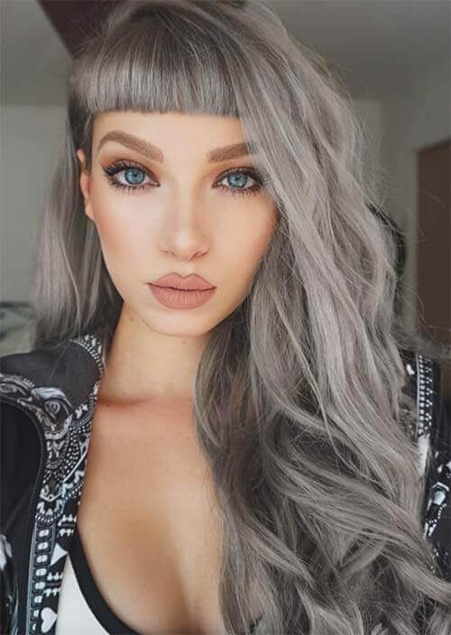 A woman's striking hairstyle featuring silvery-gray waves with subtle darker undertones, sleek straight-across bangs, and enchanting gray highlights, creating an edgy yet ethereal and dramatic chic look with sultry allure.