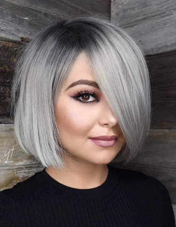 A woman's chic bob adorned with deep charcoal roots gracefully transitioning into a silvery-gray cascade, featuring subtle layers for volume and a sweeping side fringe.