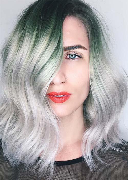 A woman's tantalizing wavy hair featuring a striking interplay of deep green roots melting into icy-gray highlights, with gently layered locks for soft, natural movement and an edgy center-parted fringe.