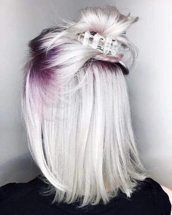 A woman's hair with an icy silver base and striking lavender roots, styled sleek and straight with soft layers. The top section is twisted into a playful half-bun, secured with a translucent clip, creating a trendy and sophisticated look blending gray tones with a refreshing touch of lavender.