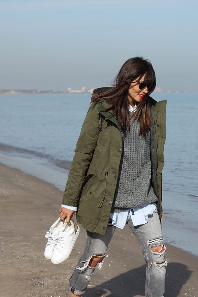 Olive green hooded parka layered over a textured grey sweater and light blue shirt, paired with distressed light-wash jeans and white sneakers.