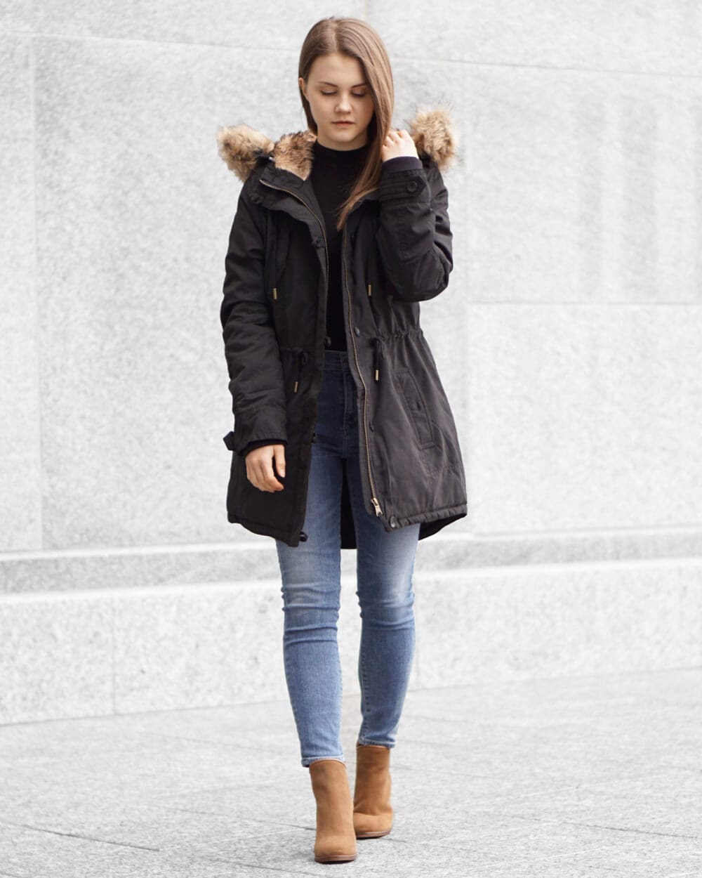 Black parka with faux fur-lined hood and cinched waist, paired with light blue skinny jeans and tan ankle boots, ideal for cold weather.