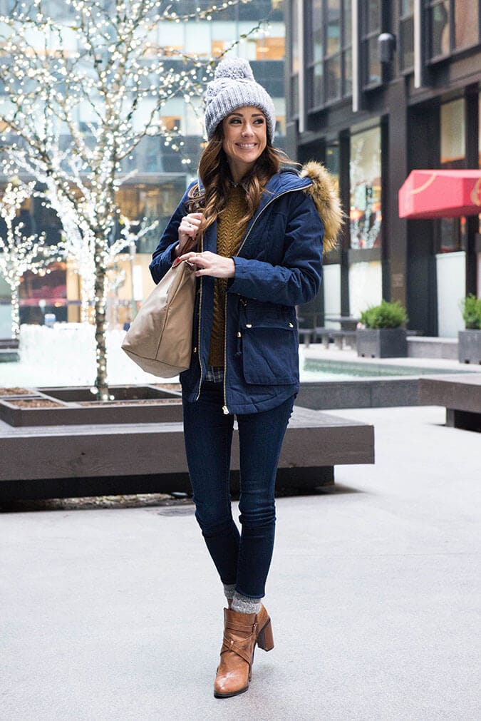 Navy parka with fur-trimmed hood, mustard sweater, dark jeans, brown ankle boots, gray beanie, and beige handbag, ideal for winter.
