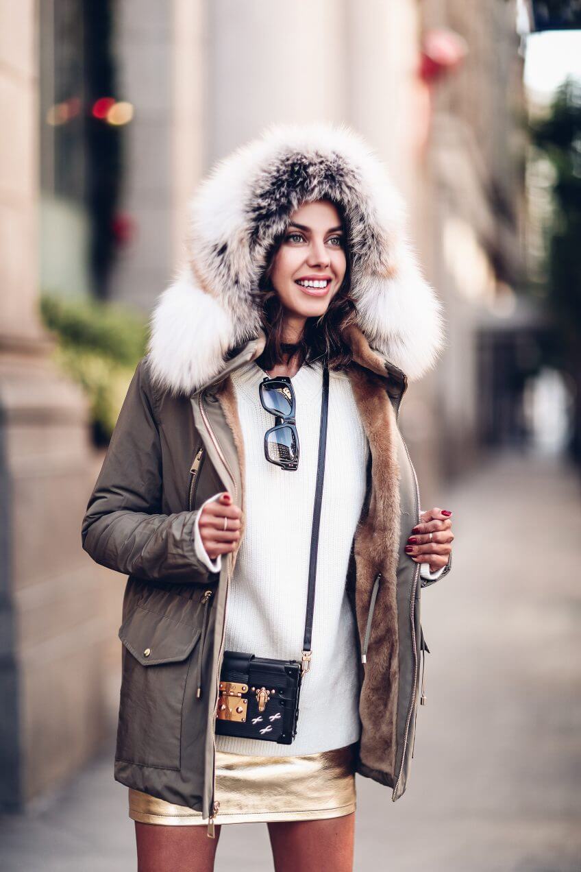 Olive green parka jacket with a fur-lined hood, layered over a cream sweater and gold mini skirt, ideal for chic winter outings.