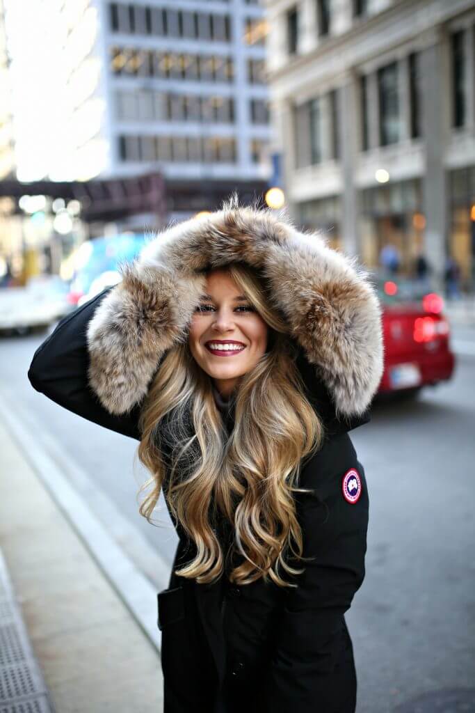 Long, flowing hairstyle peeks from beneath a fur-trimmed hood of a black parka, styled for warmth in a cozy urban winter setting.
