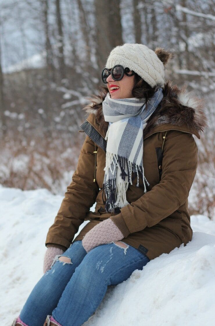 Cozy winter outfit featuring a brown parka jacket with fur-trimmed hood, white knit hat, striped scarf, and ripped blue jeans, set in a snowy landscape.