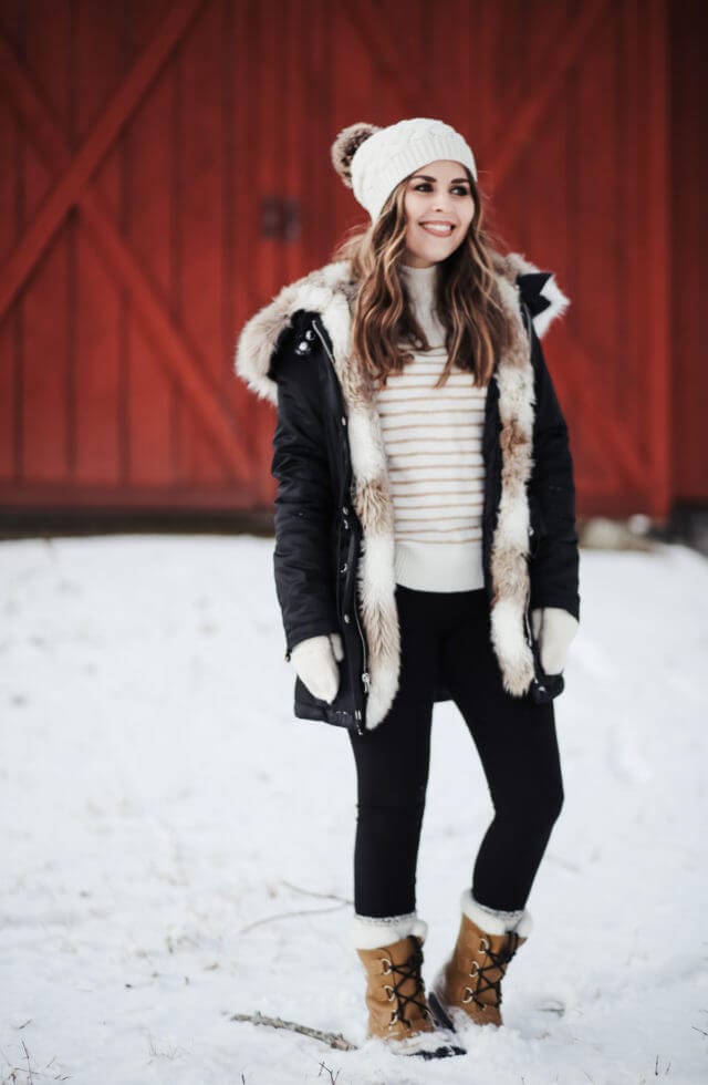 A woman stands in the snow wearing a black parka with a fur-lined hood, cream knit beanie, white striped sweater, and brown winter boots.