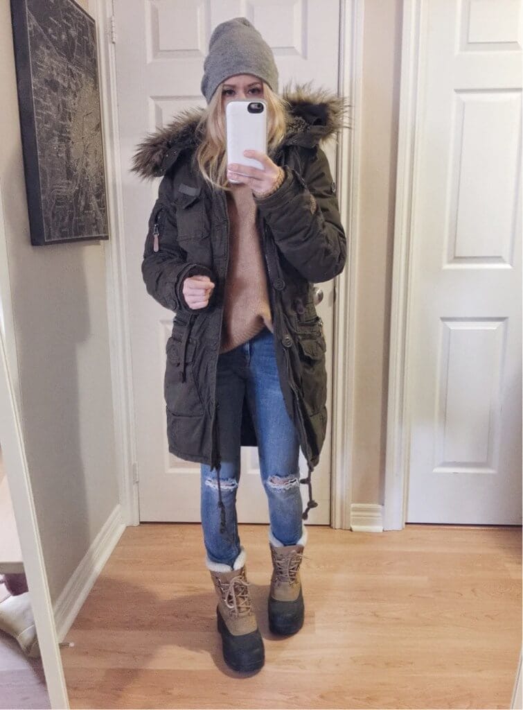 Olive green parka with faux fur hood, layered over a beige knit sweater, distressed jeans, and knee-high brown boots. Winter-ready look.