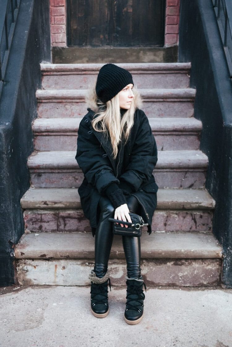 Casual winter outfit with a model on stone steps, wearing a long black parka, leggings, winter boots, and a knitted beanie.
