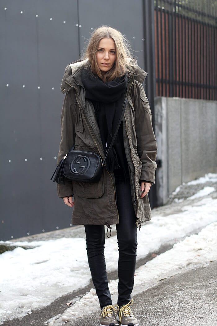 Model in an oversized olive green parka, black scarf, and fitted black pants on a snowy sidewalk, blending style and warmth.