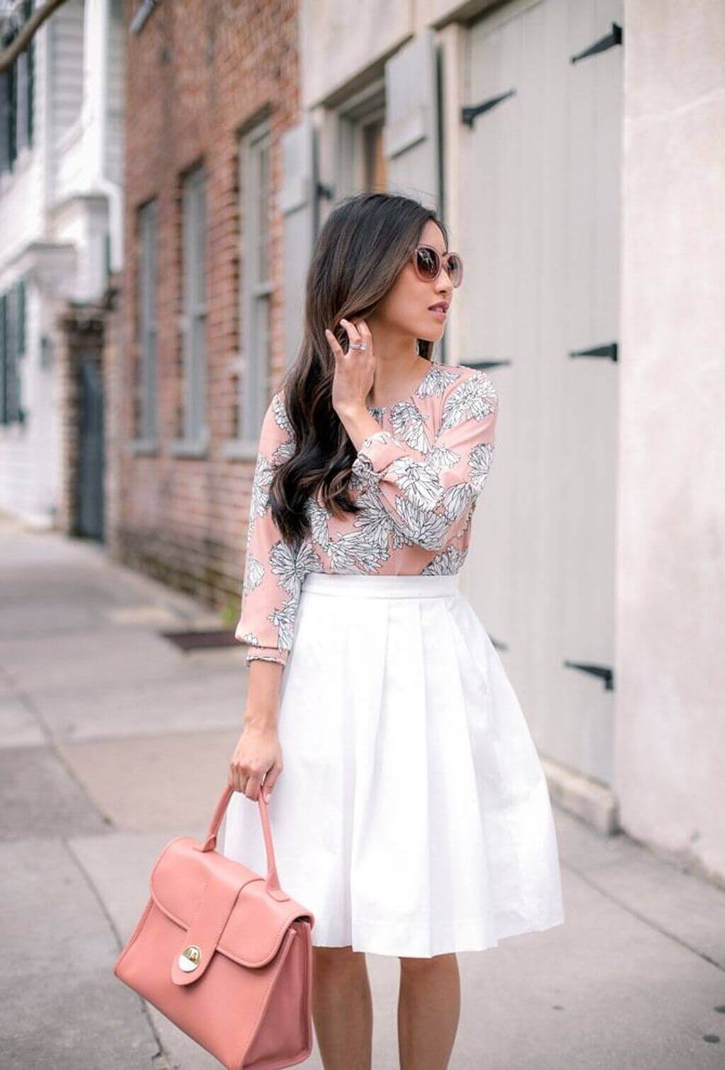 Light pink floral blouse with voluminous sleeves, white pleated skirt, oversized sunglasses, and pink handbag. Chic and feminine office attire.