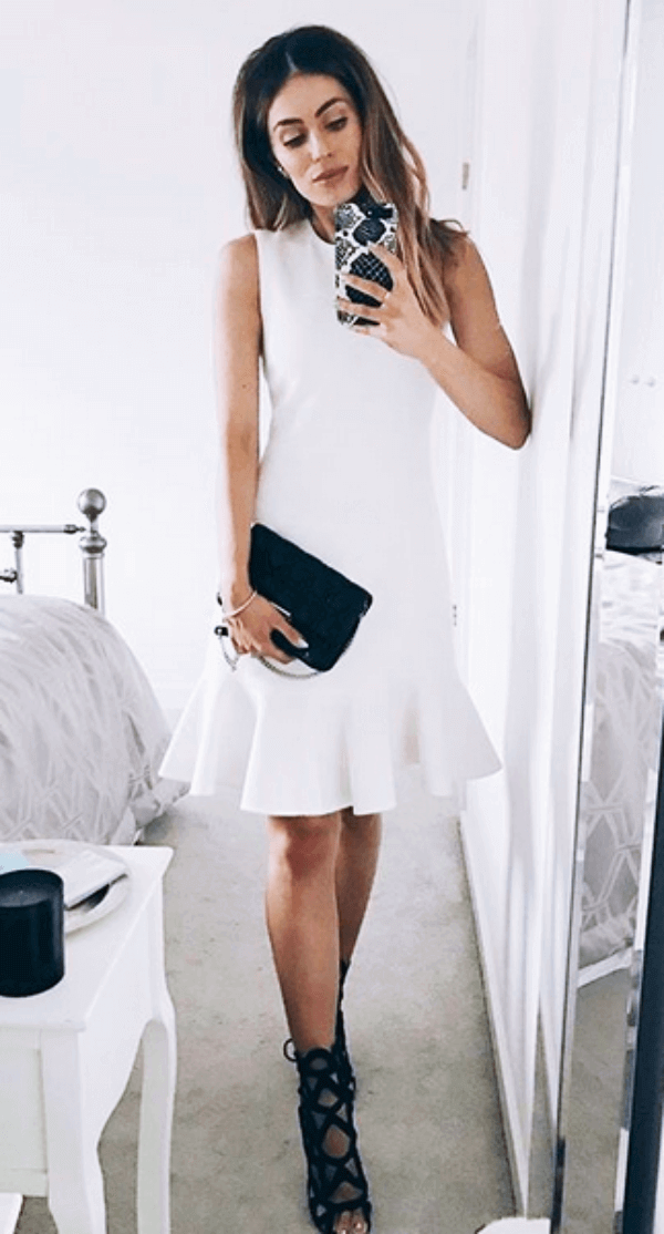 A woman in a white dress with a fitted bodice and flared hemline, paired with black strappy heels, set against light walls.