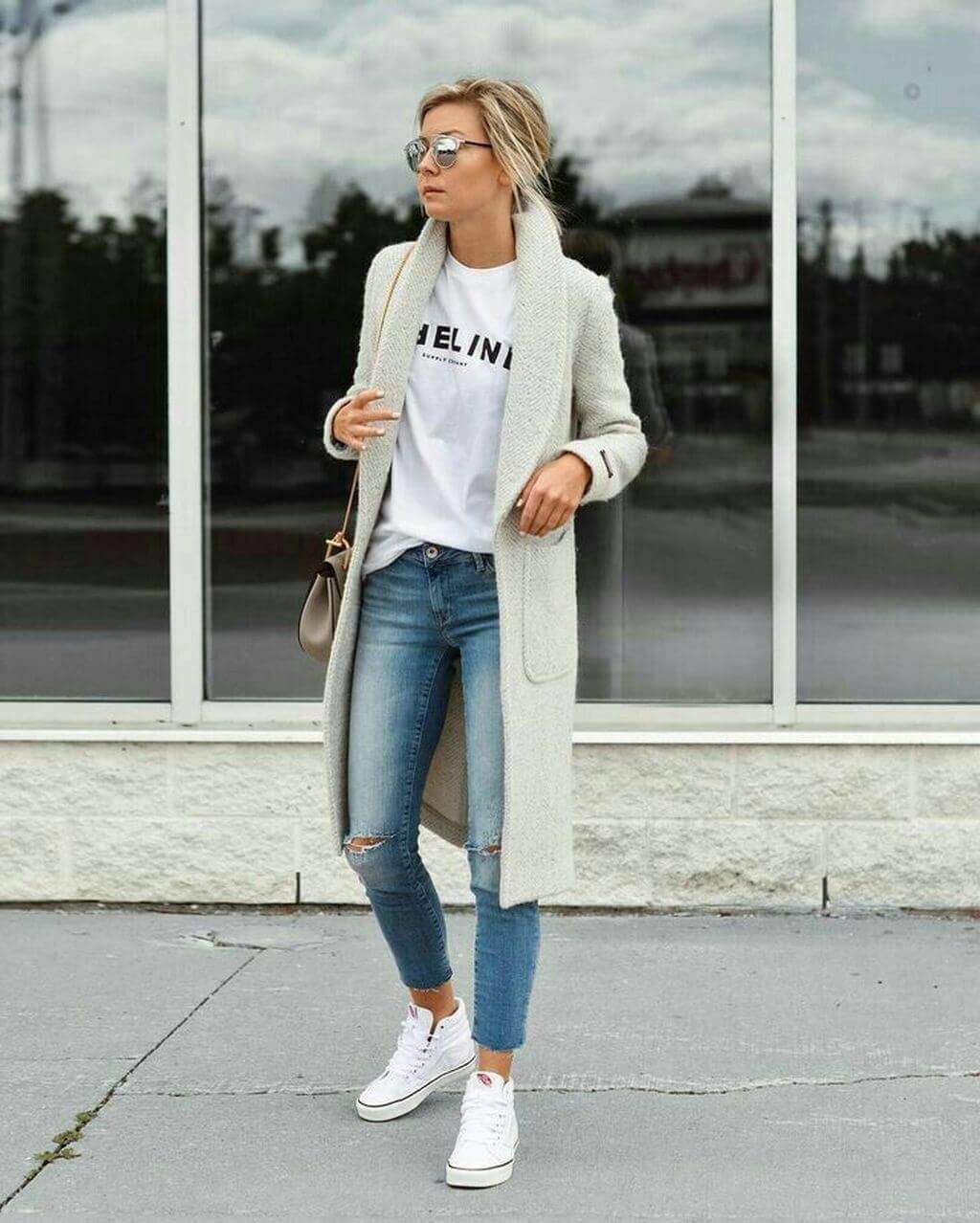 Casual office attire for women featuring a light gray cardigan over a white t-shirt, distressed light blue jeans, and white sneakers.