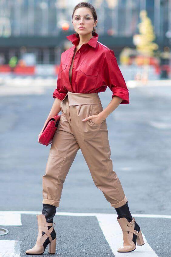 Striking red oversized blouse paired with high-waisted tan faux leather pants, accented by a vibrant red clutch and clear eyeglasses. Urban backdrop.