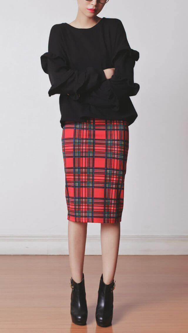 Knee-length plaid skirt in vibrant red, paired with a black long-sleeve top featuring ruffled cuffs, styled with black ankle boots.