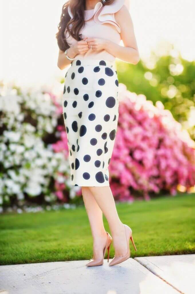Fitted midi skirt with black polka dots on white, paired with a soft pink ruffled top and nude stilettos in a vibrant garden.