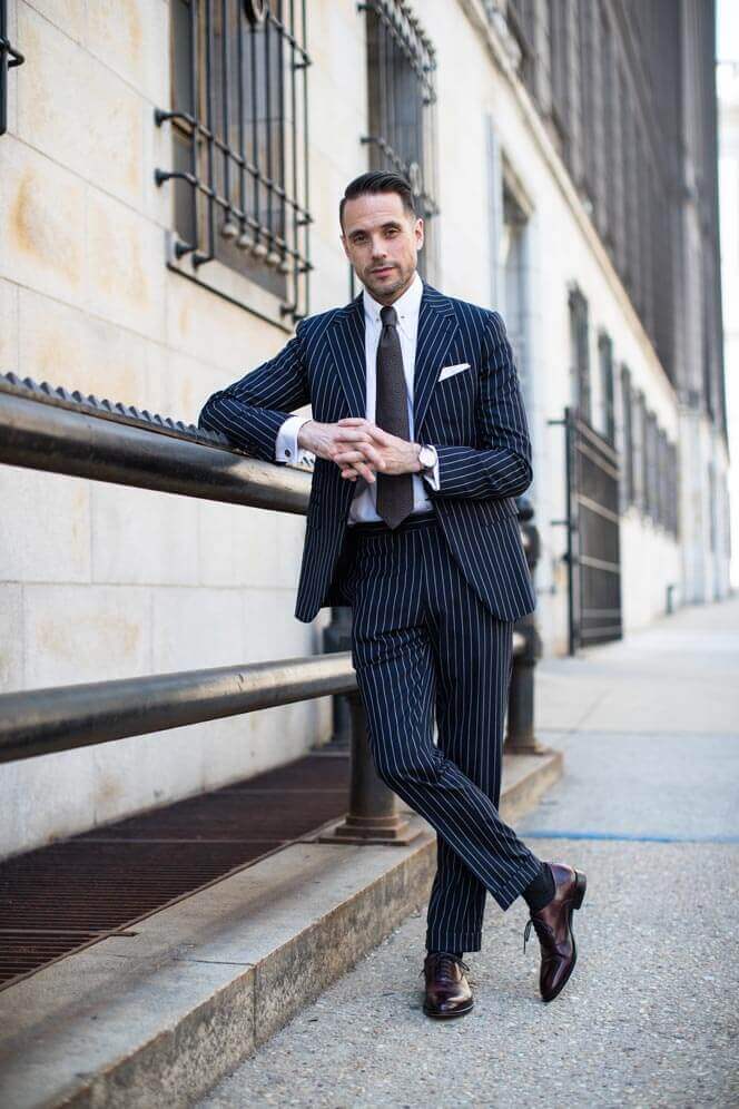 Tailored navy and white pinstripe suit with a slim lapel blazer, white dress shirt, and matching tie, paired with polished brown shoes.