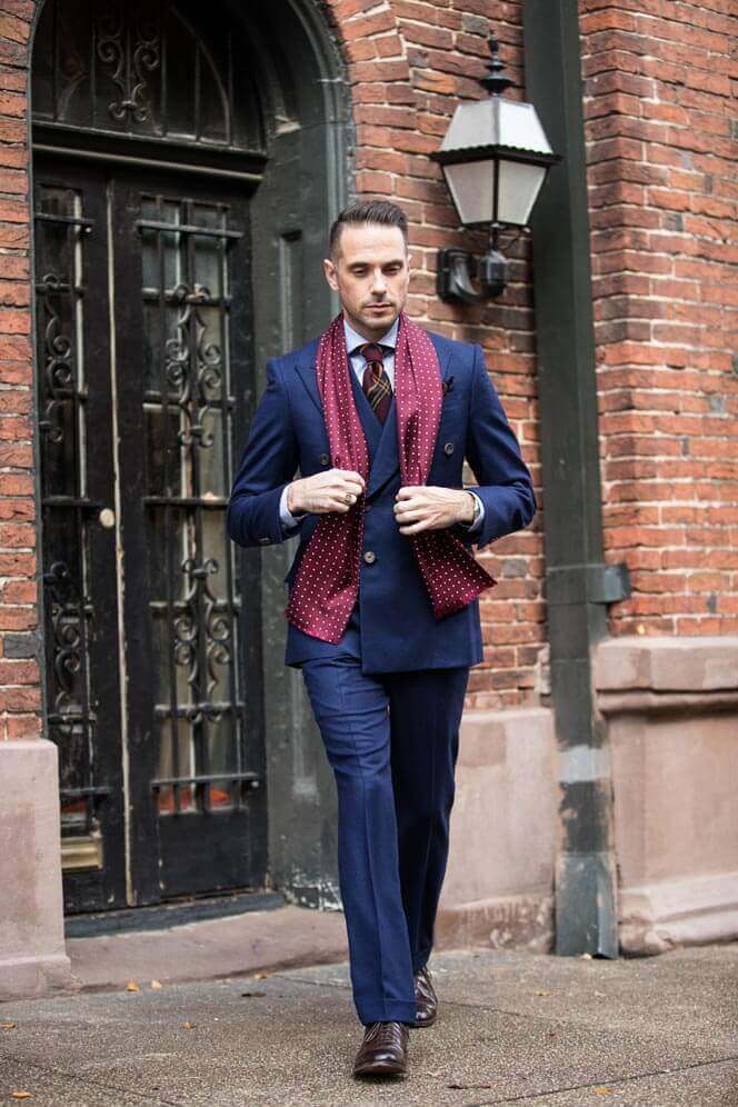 Sophisticated men's outfit featuring a tailored navy suit, fitted blazer, vest, and a burgundy polka dot scarf, walking on cobblestones.