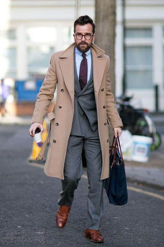 Stylish man in a tailored gray suit with a beige overcoat, brown leather shoes, holding a coffee cup and dark blue tote bag.