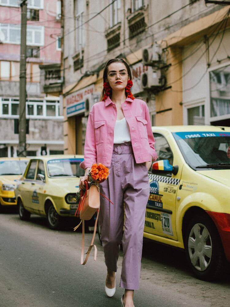 Model in pastel outfit: pink denim jacket, lavender trousers, and red floral accents. Urban street with vintage taxis, capturing soft, lively style.