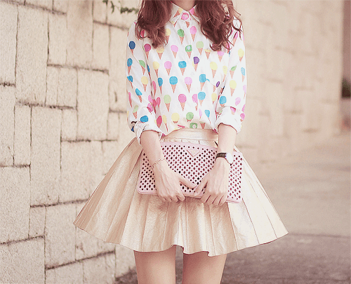 A white blouse with colorful balloon prints paired with a pleated gold mini skirt, complemented by a light pink clutch, embodying soft girl aesthetic.