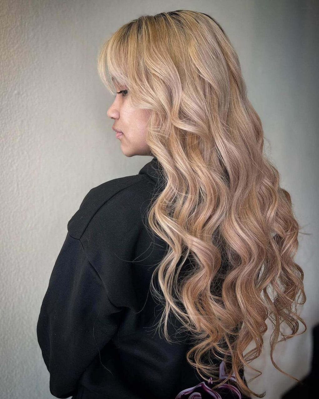 Blonde balayage with darker roots and honey-platinum highlights, styled in long layers and loose curls with a side fringe
