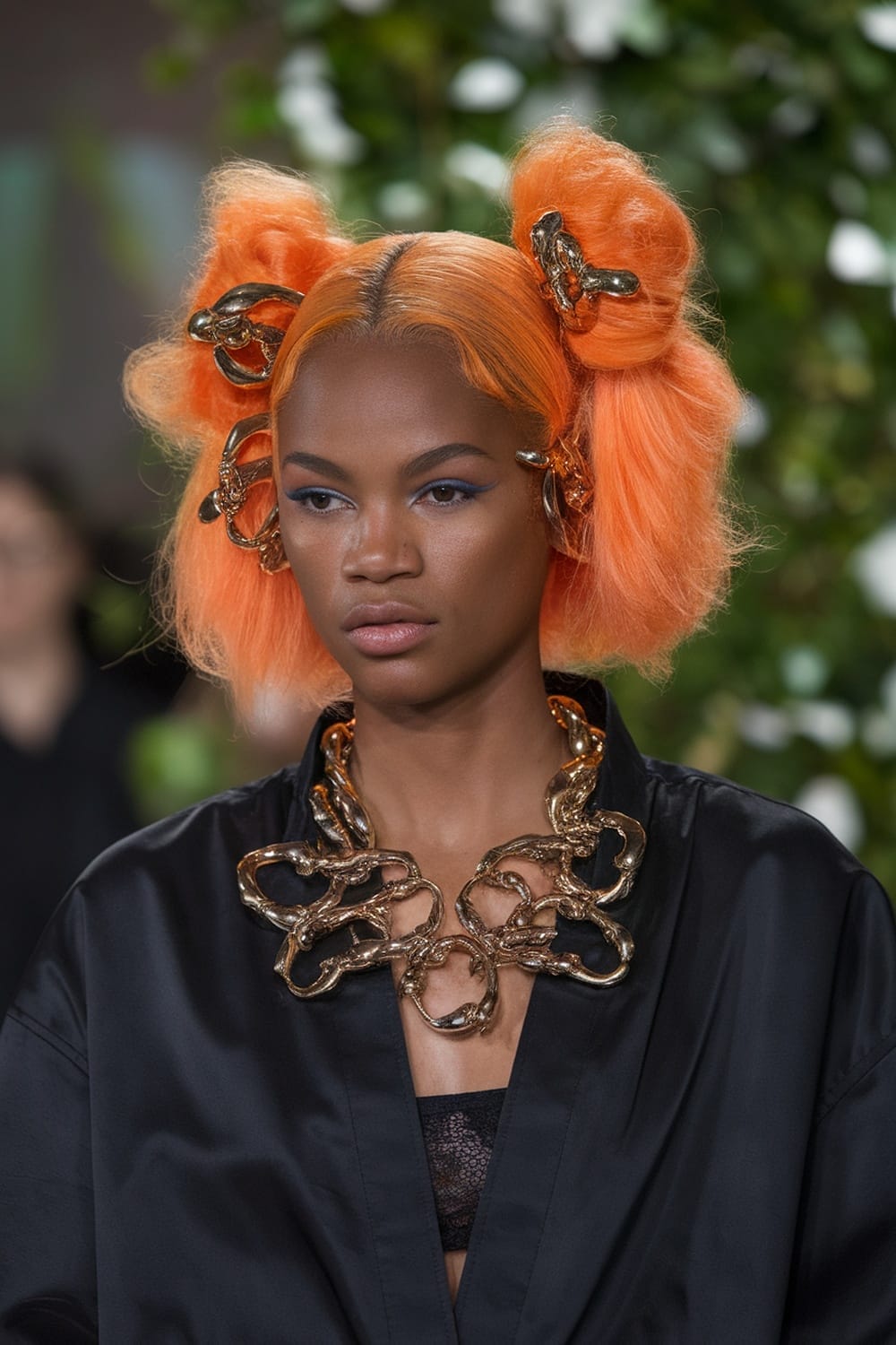 A model with orange hair styled in buns, wearing a black outfit and gold accessories.