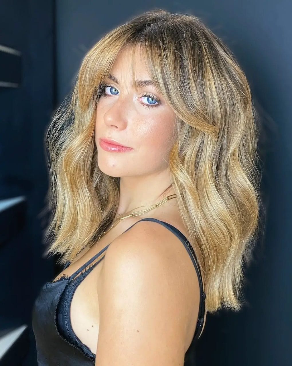 Balayage blending of blonde and brunette hues with curtain Bardot bangs, showcasing modern grace with classic charm.