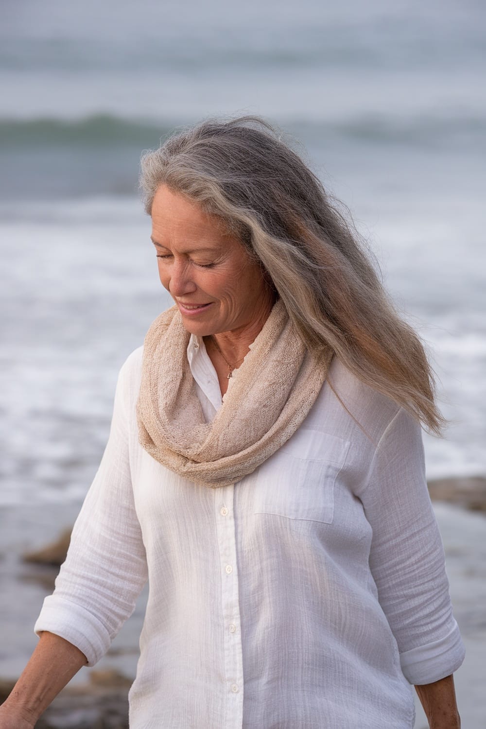 A woman with long beachy textured hair, wearing a light-colored scarf and a white shirt, standing by the beach.