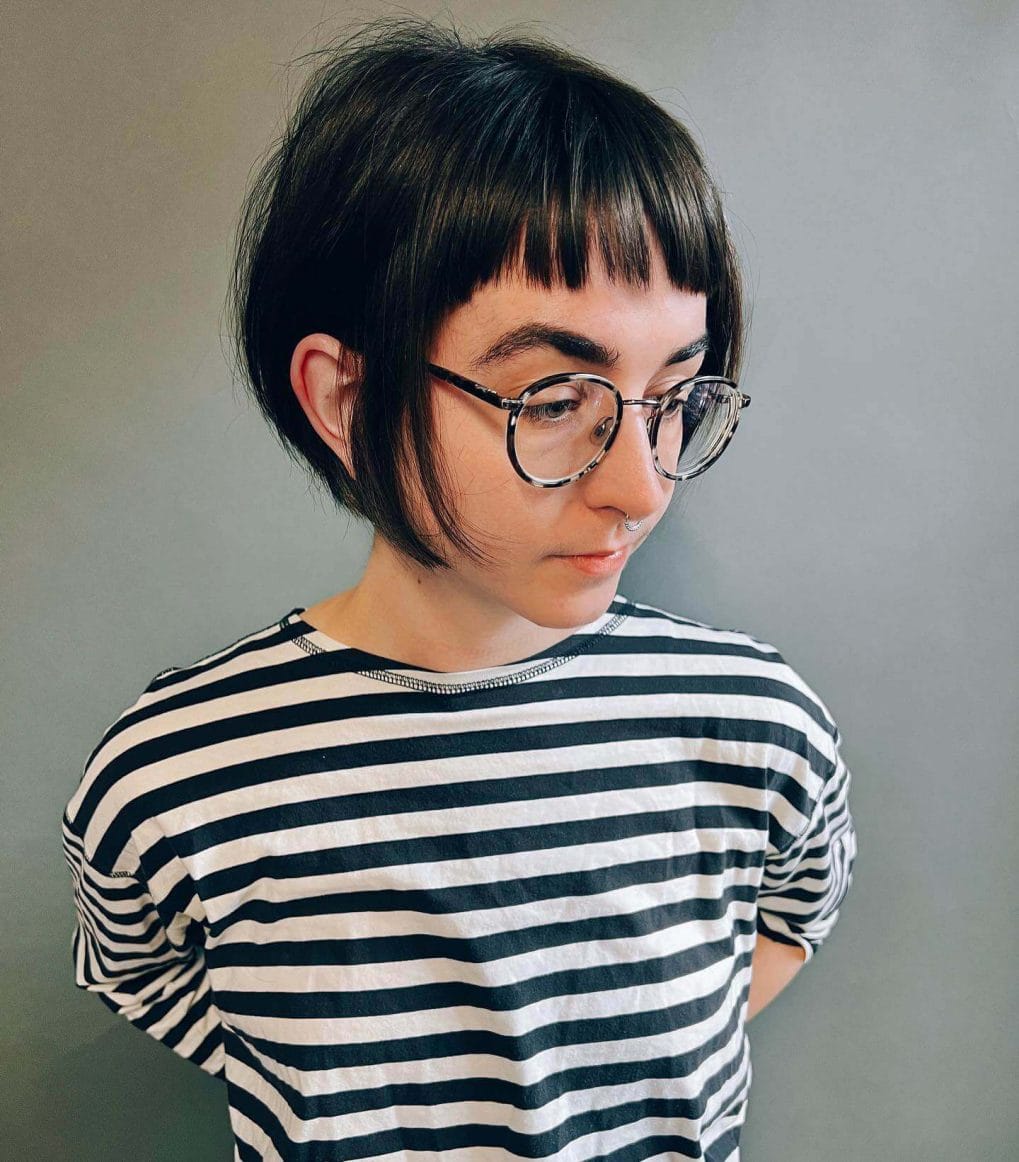 Black pixie cut with full straight fringe and circular glasses frames.