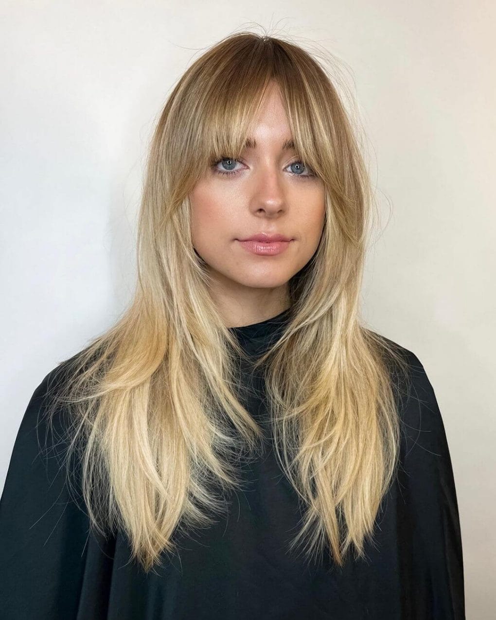 Long layers in a gradient of blonde hues with bold full bangs, presenting a sophisticated and sharp look.