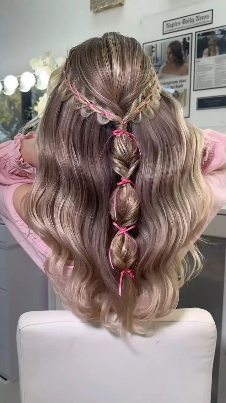 Half-up, half-down hairstyle with a braided crown, pink ribbons, and balayage-highlighted waves.