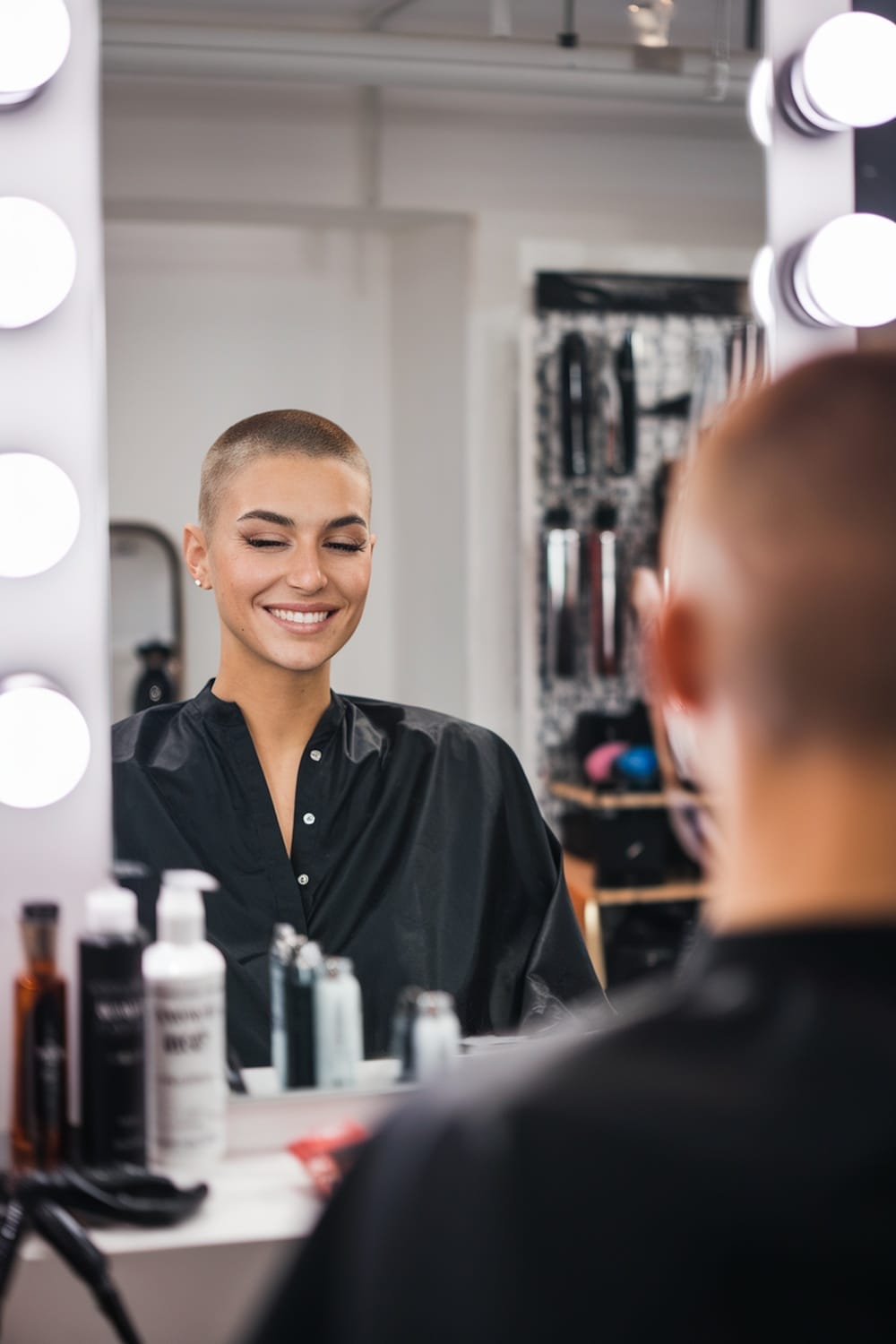 A woman with a buzz cut smiling in front of a mirror at a salon.