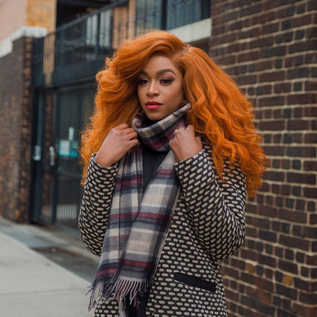 A confident black woman with ginger hair, dressed in a patterned coat and scarf, standing against a brick wall.