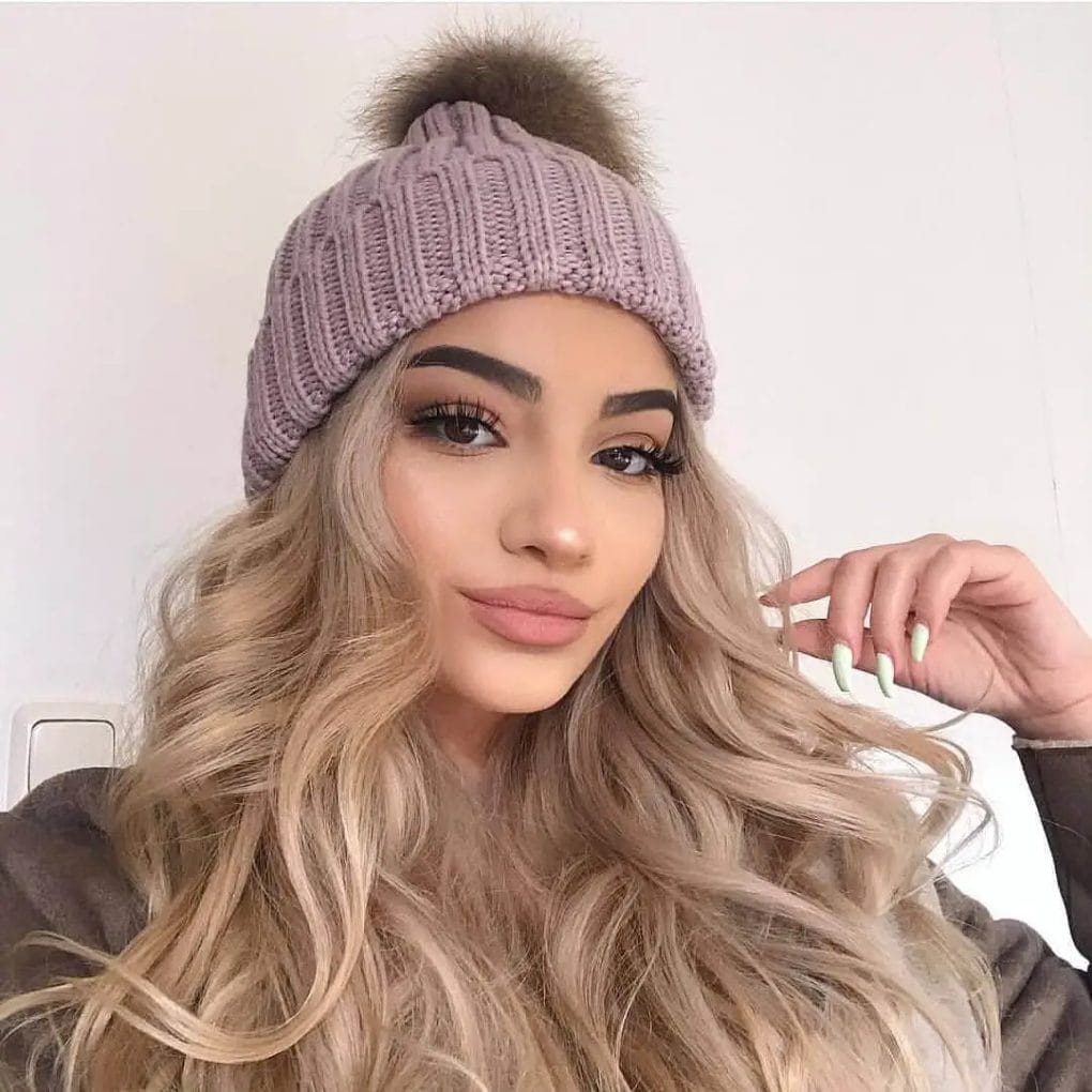 Luxurious honey-blonde waves spilling from beneath a pastel beanie with pompom.