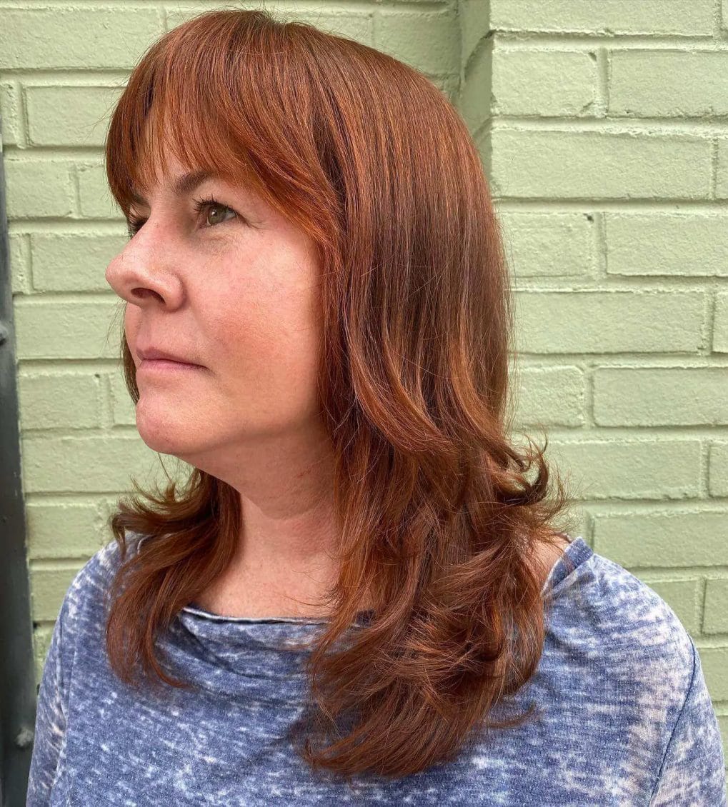 Layered haircut with rich auburn tones, straight-across bangs, and subtle feathering.