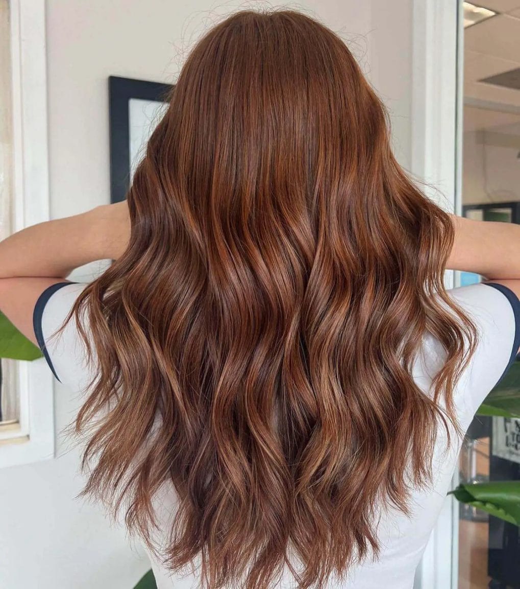 Long mane with brunette to copper gradient, polished balayage, and cascading waves.