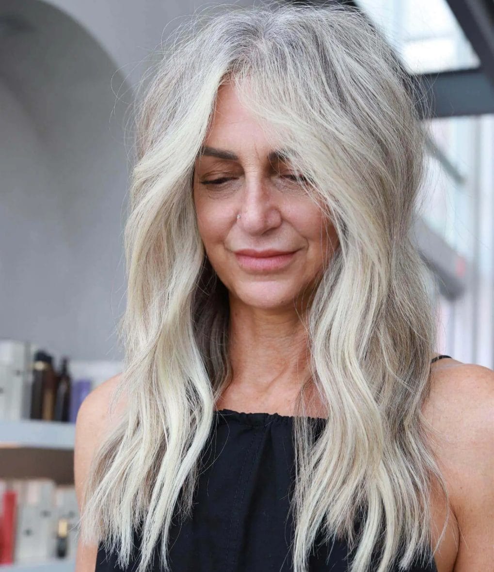 Long layered hairstyle with natural waves, silver to blonde ombre, and subtle off-center parting.