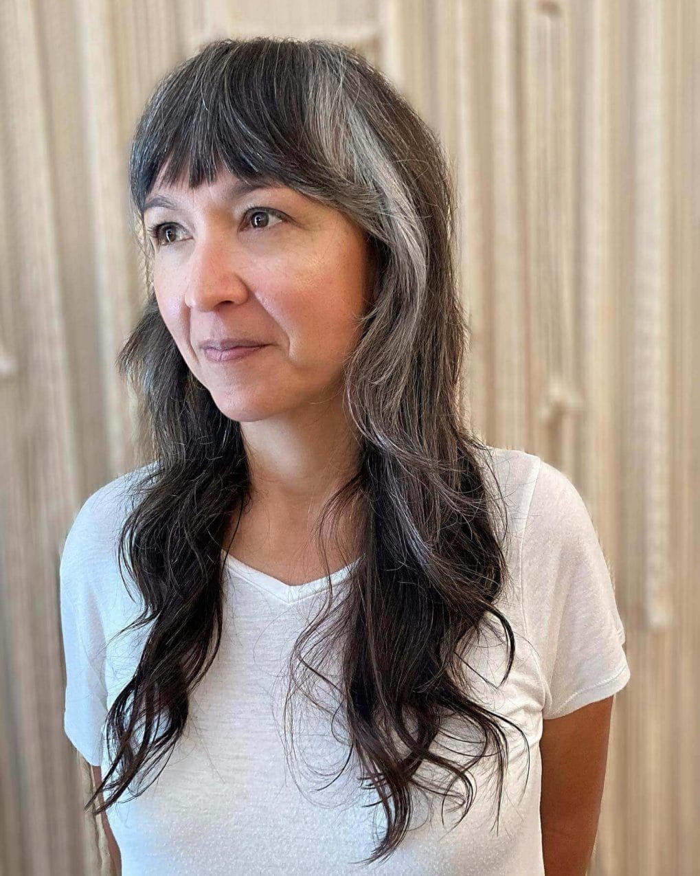 Long flowing layers with relaxed waves, dark hair with gray highlights, and full straight-across bangs.