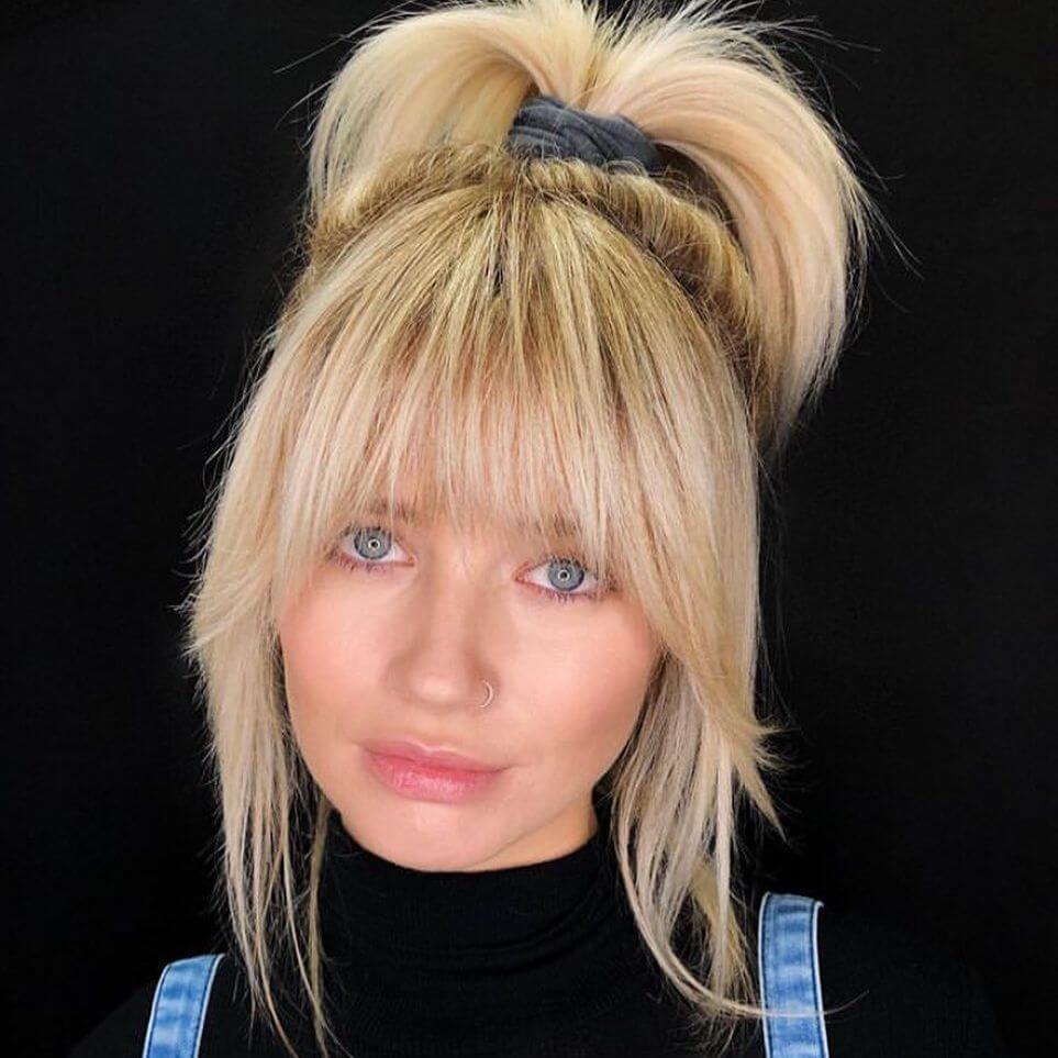 Playful high ponytail in blonde with voluminous texture and full eyebrow-skimming bangs, adding a youthful touch.