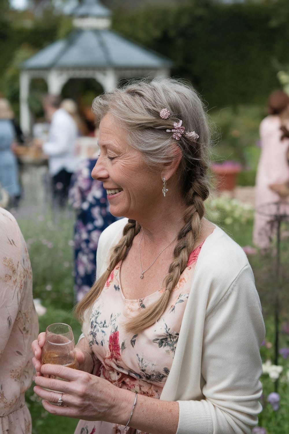 A woman with loose braids and floral hair accessories, smiling while holding a drink in a garden setting.