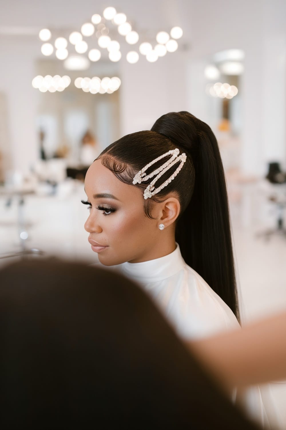 A woman with a sleek ponytail hairstyle adorned with pearl accessories in a bright salon setting.