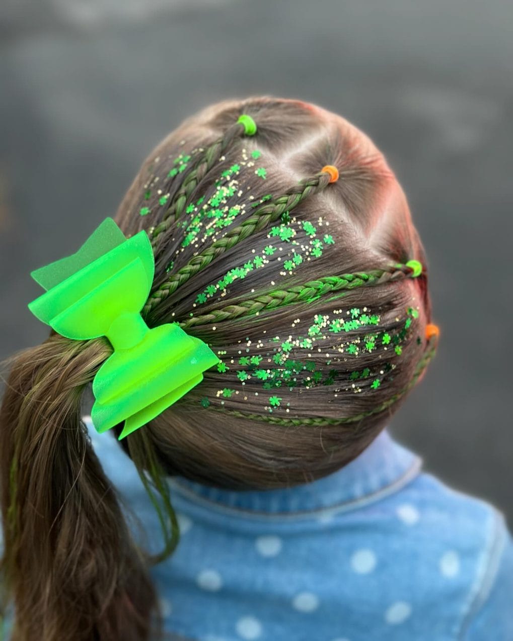 Festive gymnastics hairstyle featuring small braids with green ties and glitter, topped with a vibrant bow