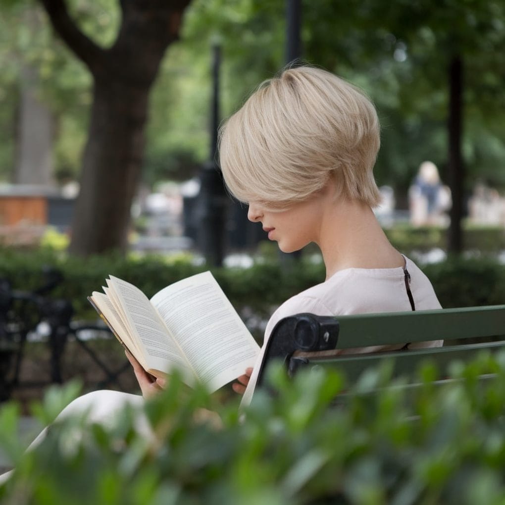 A woman with a soft boyish haircut reading a book in a park.