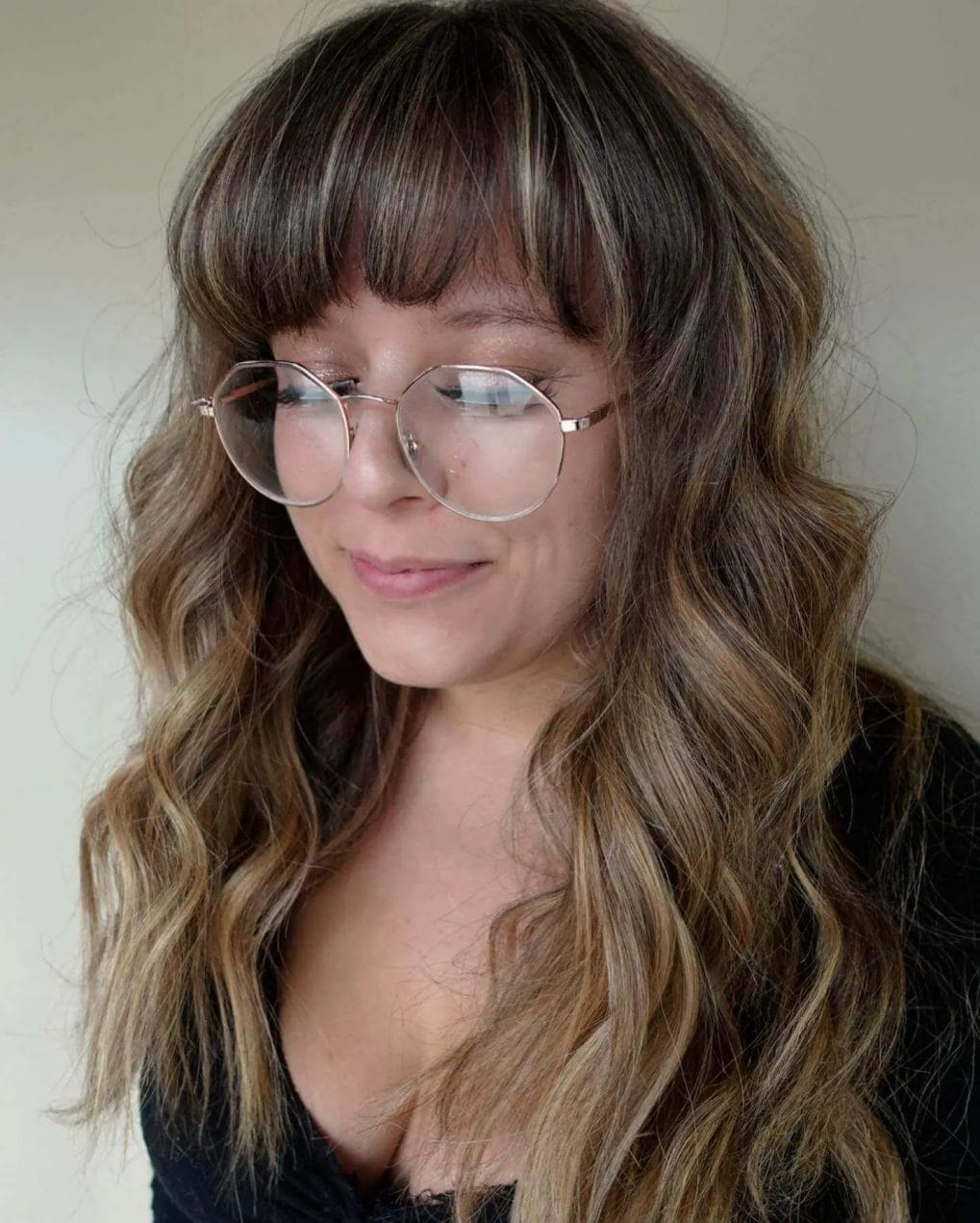Sun-kissed highlights in chic wavy hair with full, straight fringe and round spectacles.